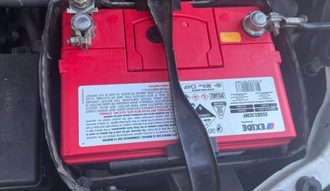 Close-up of a red car battery with terminal connectors and warning labels visible inside the engine compartment