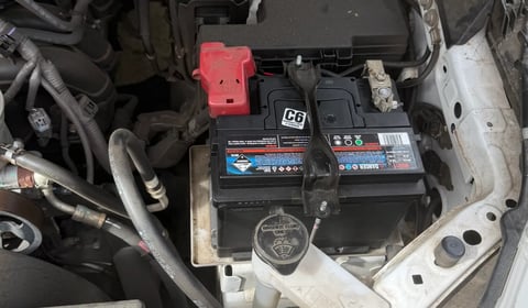 Car engine bay showing two batteries, red positive terminal clamp, and various hoses and wiring under the hood