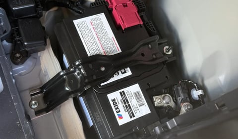 Car engine compartment showing battery with red positive terminal and printed specification labels on black components