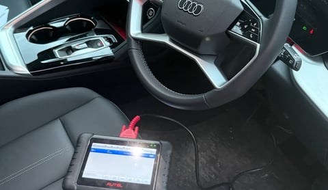 Car interior showing Audi steering wheel, dashboard, and diagnostic scanning device on floor mat